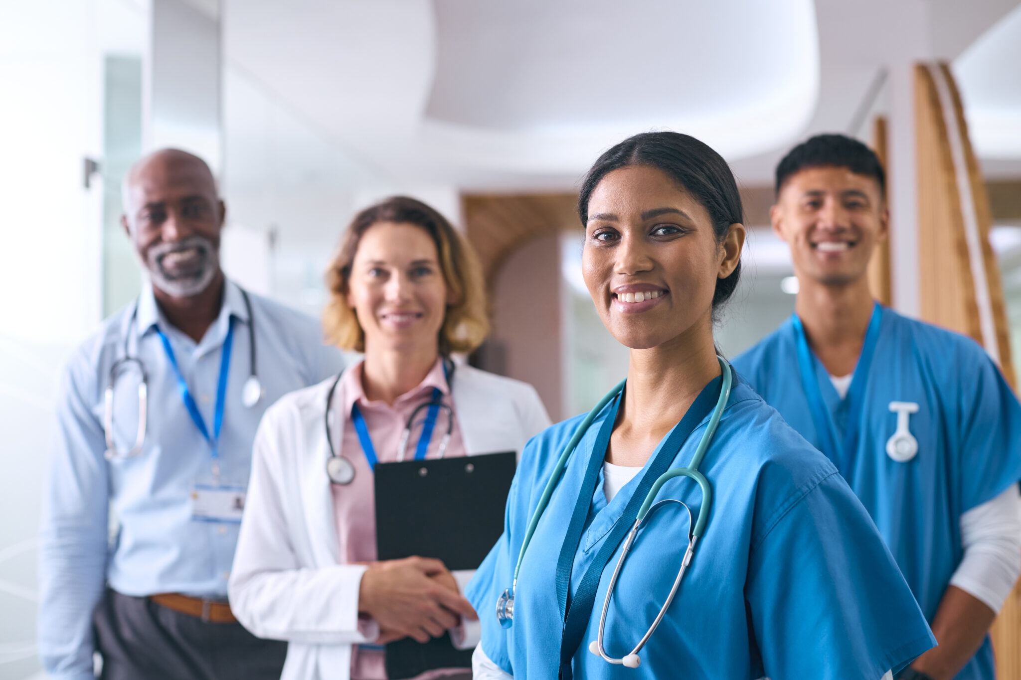 Portrait Of Smiling Multi Cultural Medical Team Wearing Scrubs And White Coats In Modern Hospital